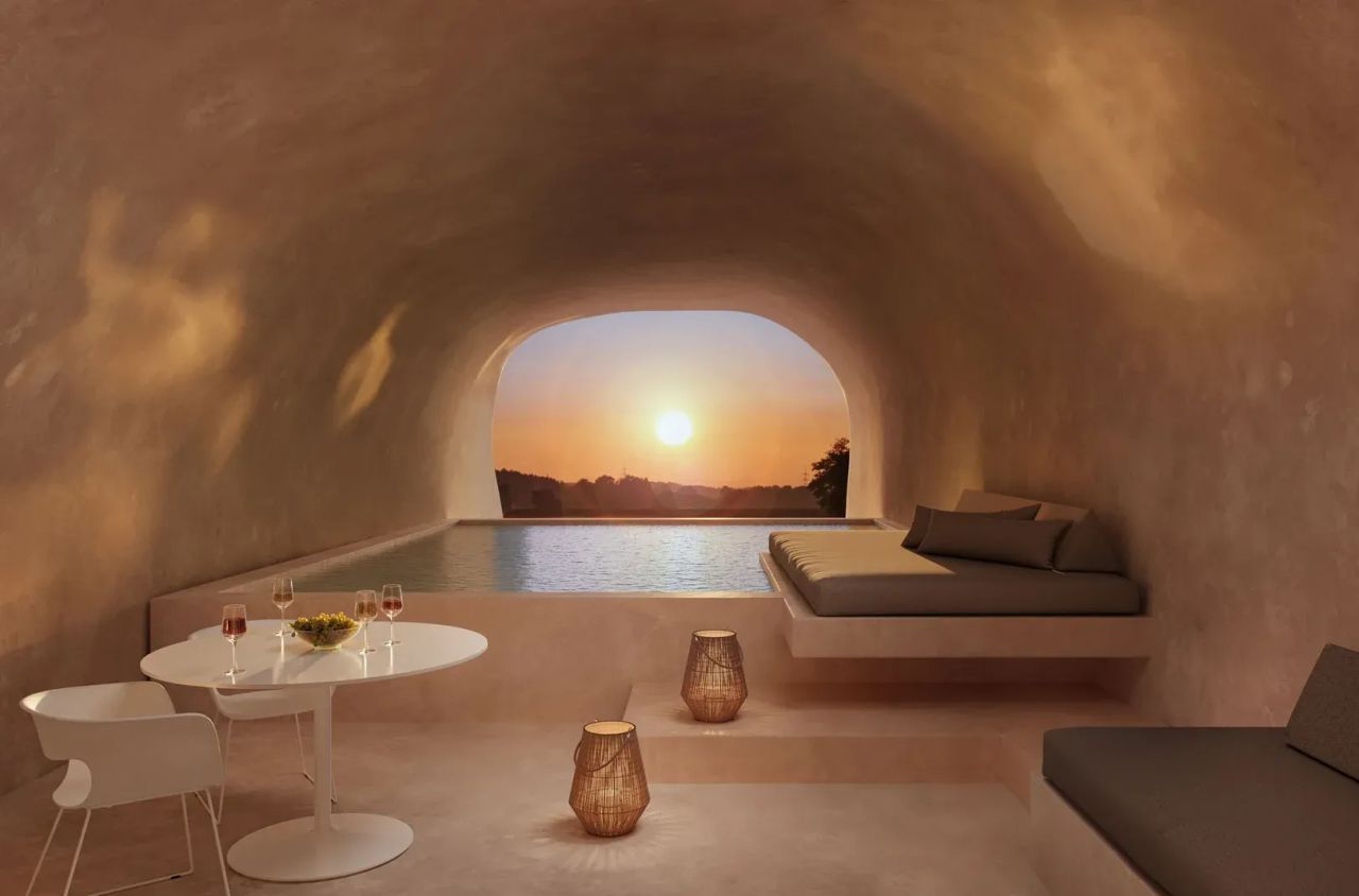 Product rendering showing a cave-style room with an indoor pool, lounge beds, a round table with chairs, and a sunset view